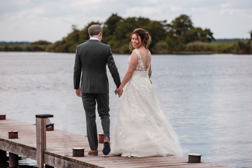 Bruidspaar op een steiger, hand in hand. Bruid kijkt achterom naar fotograaf